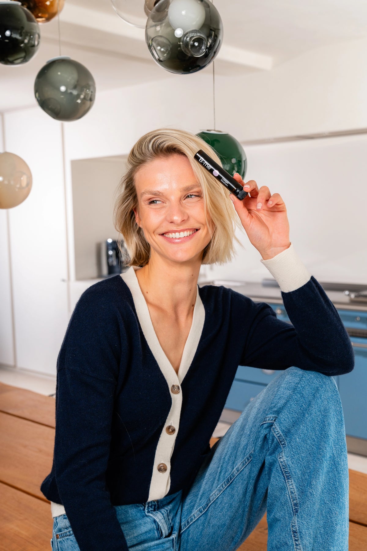 Vrouw glimlacht terwijl ze B.me Collageen Advance+ shot vasthoudt in haar keuken – supplement voor huidverzorging van binnenuit.