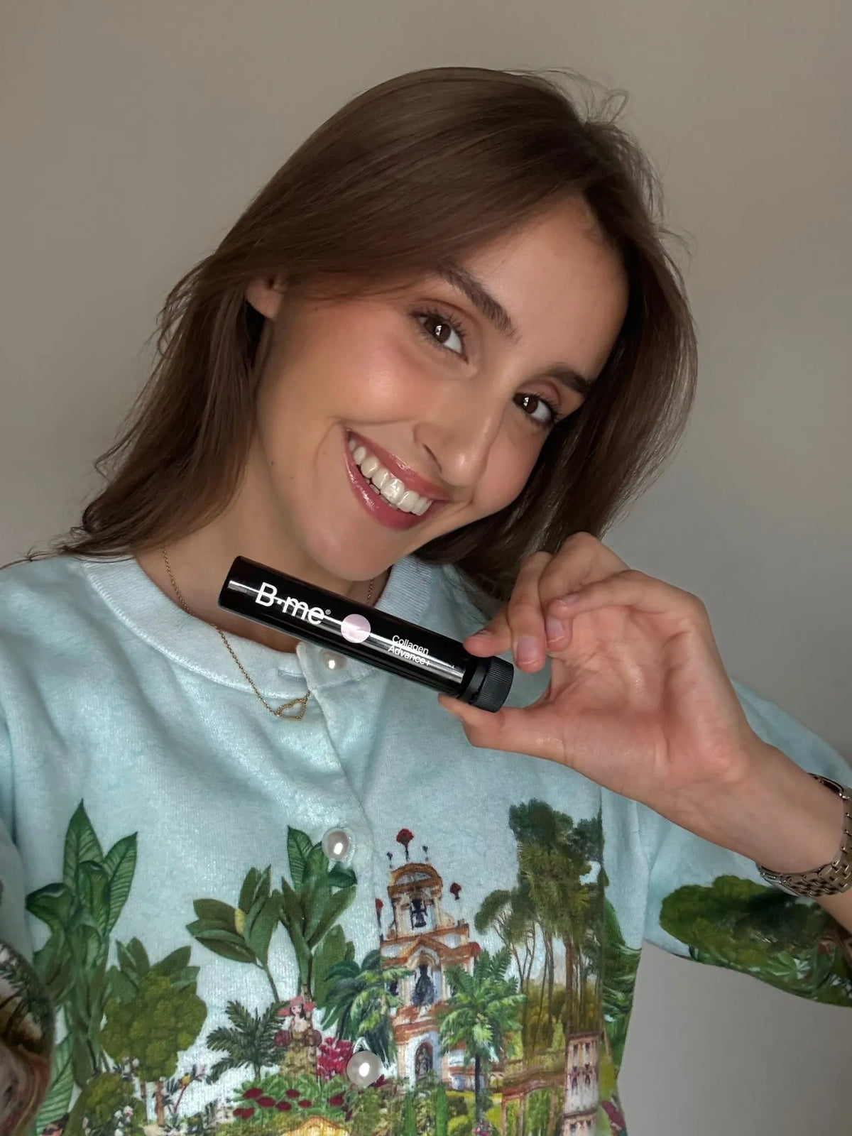 Kenza smiling and holding a B.me Collageen Advance+ shot, featuring Peptan collagen, Bloosil, and vitamin C, wearing a light blue shirt with a colorful print.